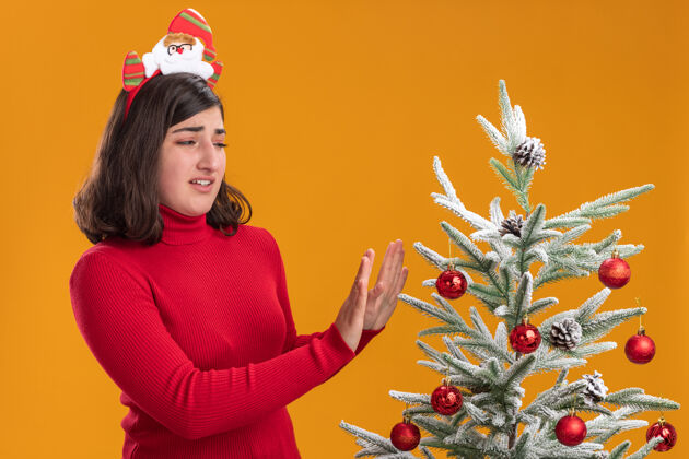 手穿着圣诞毛衣的年轻女孩戴着有趣的头带 站在橙色背景下的圣诞树旁看头带恶心