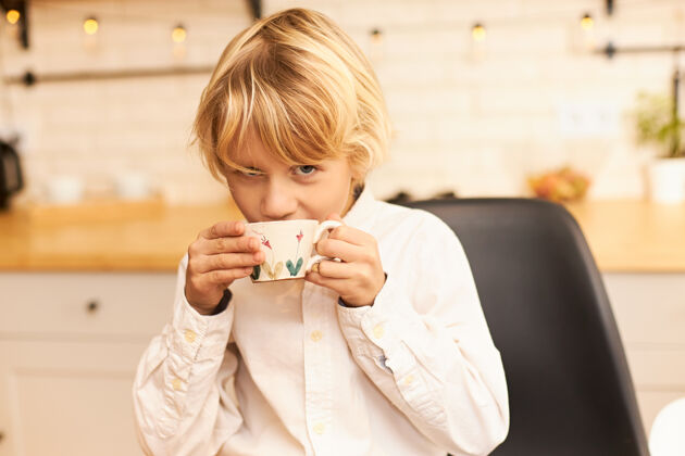 小一幅美丽快乐的金发男孩的画像 在放学前一边喝茶一边吃早餐 手里拿着杯子 微笑着拿着厨房柜台上的餐具和花环果汁杯子孩子