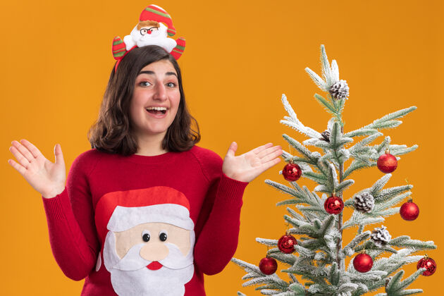 微笑穿着圣诞毛衣 戴着滑稽头带 脸上带着微笑的快乐小女孩站在橙色墙上的圣诞树旁毛衣头带戴着