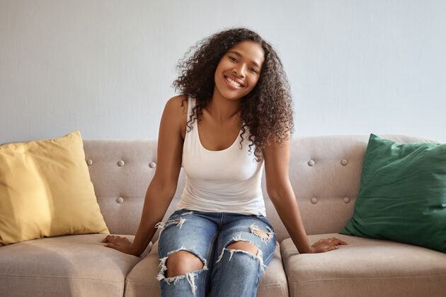 女人人们和生活方式穿着蓝色破洞牛仔裤和白色背心的年轻漂亮黑皮肤女人在家里放松 坐在舒适的带靠垫的灰色沙发上 面带微笑休闲舒适沙发