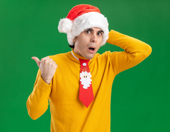帽子一个穿着黄色高领毛衣 戴着圣诞帽 打着滑稽领带的年轻人看着摄像机 惊讶地指着后面站在绿色背景上的镜头圣诞老人背部领带