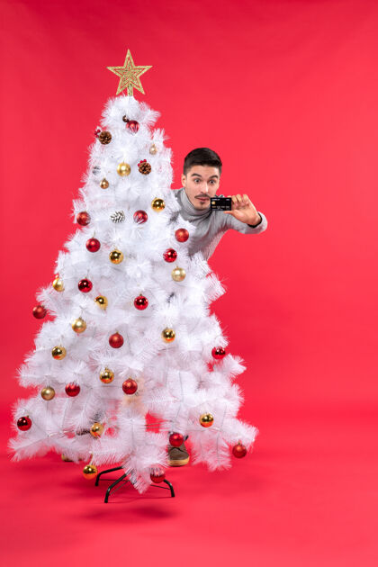 庆祝一个穿着灰色上衣的年轻人站在装饰过的圣诞树后面 看着红色的手机成人年轻人圣诞节