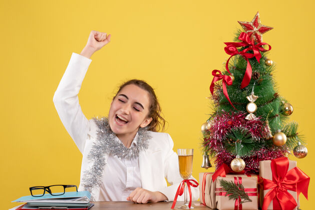 前面正面图：女医生坐在桌子后面 在黄色的背景上 拿着圣诞树和礼品盒 欢呼雀跃快乐桌子高兴