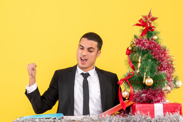 礼物前视图满意的男子坐在桌旁 展示致胜手势圣诞树和礼物满意桌子行政人员