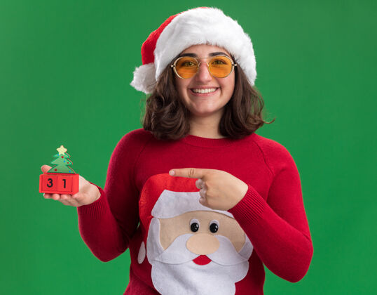 高兴穿着圣诞毛衣 戴着圣诞帽 戴着圣诞眼镜的小女孩手里拿着玩具方块 用食指指着玩具方块 站在绿色的墙上开心地笑着索引手指圣诞老人