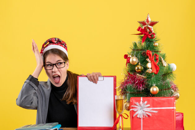装饰品前视图高喊着戴圣诞帽的女孩坐在桌边圣诞树和礼物鸡尾酒节日舞会树
