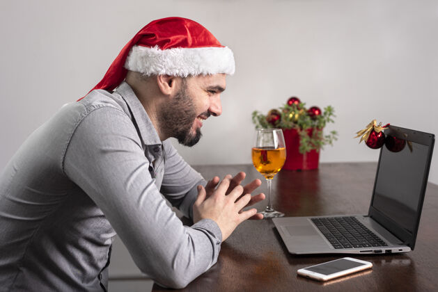 桌子特写一个西班牙裔男子戴着圣诞老人的帽子 享受他的酒 并有一个在线会议圣诞快乐圣诞会议