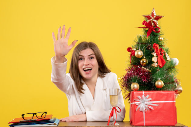 圣诞节正面图：女医生坐在黄色背景的桌子前 放着圣诞树和礼品盒办公室前面花