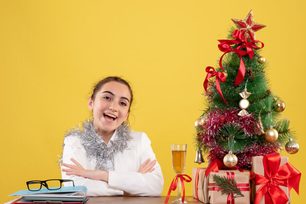 情感正面图：女医生坐在黄色背景的桌子后面 还有圣诞树和礼品盒圣诞节花家