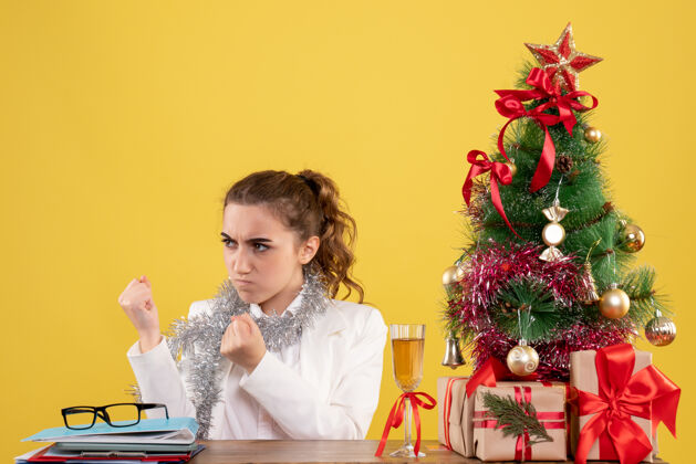 健康正面图：女医生坐在桌子后面 怒容满面 背景是黄色的圣诞树和礼盒插花情绪女医生