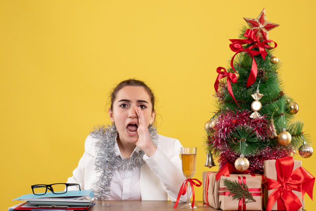呼叫前视图：女医生坐在桌子后面 用圣诞树和礼品盒在黄色背景上呼唤插花女前面