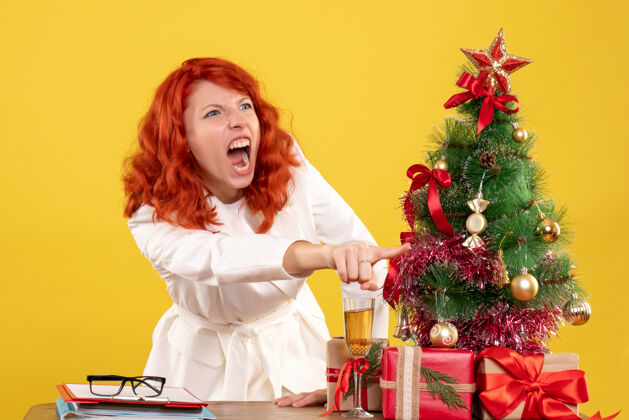 女性前视图：女医生坐在桌子后面 黄色背景上的圣诞礼物尖叫着背景前面插花