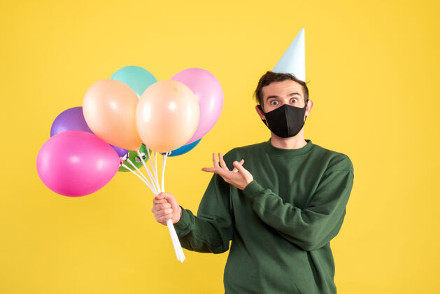 年轻正面图：戴着派对帽和黑色面具的年轻人指着黄色的气球人面具年轻人