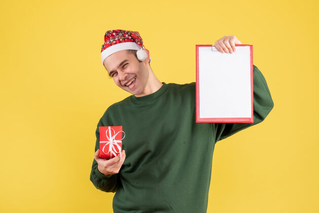 站着正面图：穿着绿色毛衣的笑脸男人拿着剪贴板和礼物站在黄色的椅子上帽子剪贴板大笑