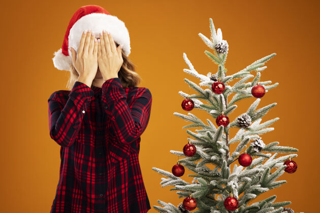 戴着快乐的年轻漂亮女孩站在圣诞树旁 戴着圣诞帽 脸上蒙着圣诞帽 手孤立在橙色的墙上快乐帽子就在附近