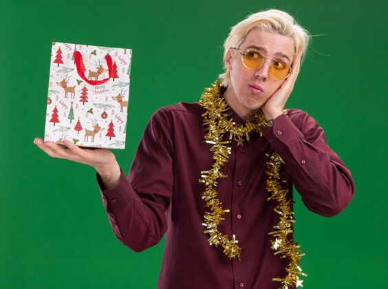 圣诞节困惑的年轻金发男子戴着眼镜 脖子上戴着金属丝花环 手里拿着圣诞礼品袋 一边看着一边把手放在脸上 隔离在绿色的墙上年轻脖子花环