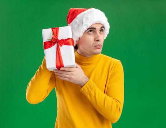 年轻人一个穿着黄色高领毛衣 戴着圣诞老人帽的年轻人站在绿色的背景上 耳边挂着一件礼物 看上去很好奇看着礼物站着