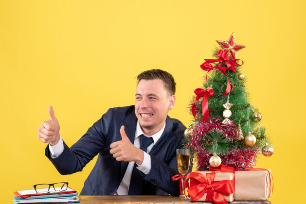 微笑正面图微笑的男人正在做竖起大拇指的标志 坐在圣诞树旁边的桌子上 黄色的礼物桌子行政人员黄色
