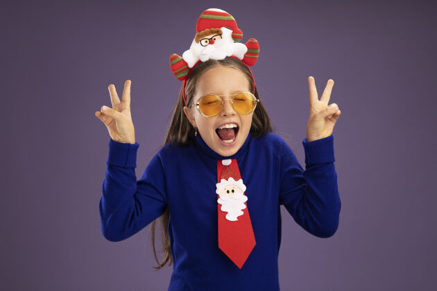 搞笑小女孩穿着蓝色高领毛衣 打着红色领带 头上戴着有趣的圣诞戒指 高兴而兴奋地在紫色背景上展示着v字标志站着Vsign圣诞节