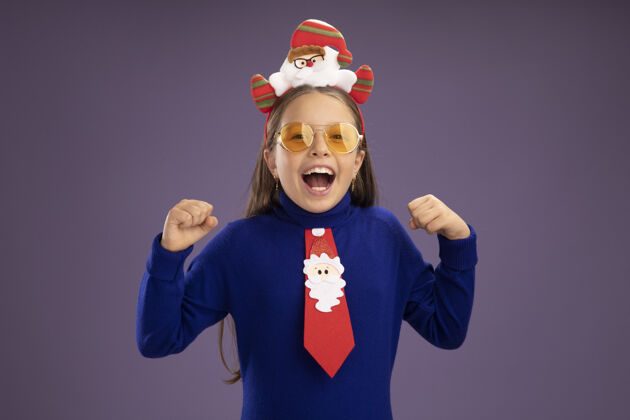 女孩小女孩穿着蓝色高领毛衣 打着红色领带 头上戴着有趣的圣诞戒指 紧握着拳头 站在紫色的背景上兴奋地尖叫着高领毛衣兴奋拳头