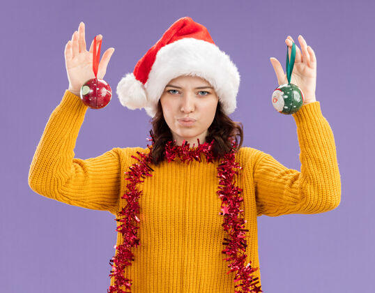 帽子自信的年轻斯拉夫女孩 戴着圣诞帽 脖子上戴着花环 手拿着玻璃球饰物 背景是紫色 有复制空间举行女孩圣诞老人