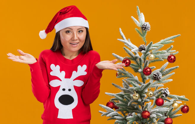 女人惊讶的年轻亚洲女孩戴着圣诞帽和毛衣站在圣诞树旁摊开手孤立在橙色的墙上附近惊讶穿着