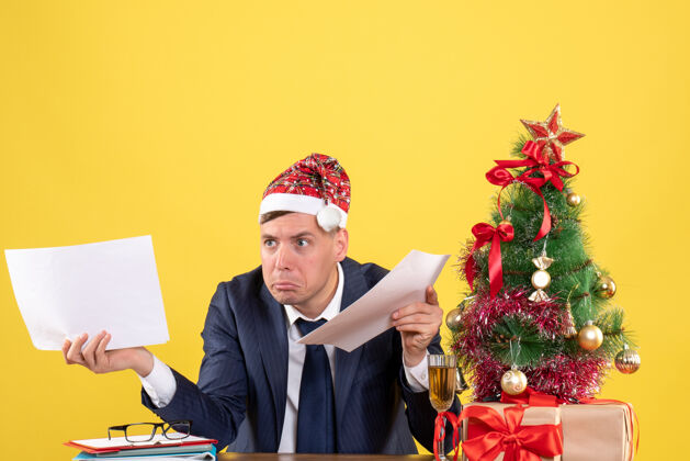 黄色前视图混乱的人拿着文件坐在圣诞树附近的桌子上 黄色的礼物坐着男人桌子