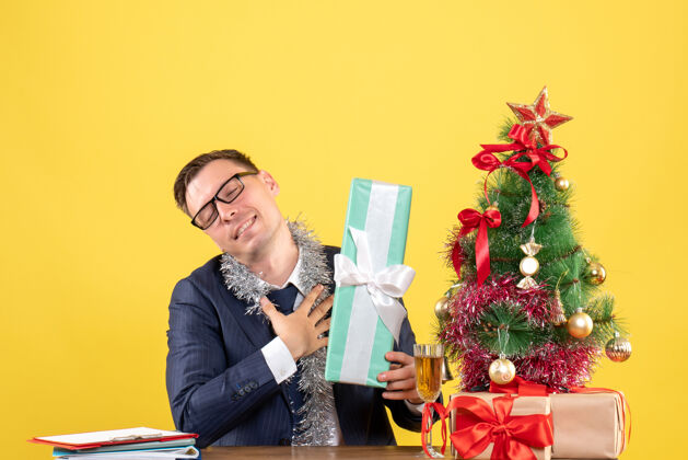 坐着满意的男人闭上眼睛坐在圣诞树旁的桌子前 黄色的礼物礼物新郎人