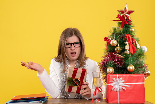 抱正面图：女医生坐在桌子前 手里拿着黄色背景的礼物 还有圣诞树和礼品盒现在微笑情感