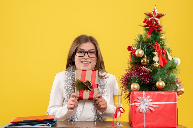 人正面图：女医生坐在桌子前 手里拿着黄色背景的礼物 还有圣诞树和礼品盒人圣诞节桌子