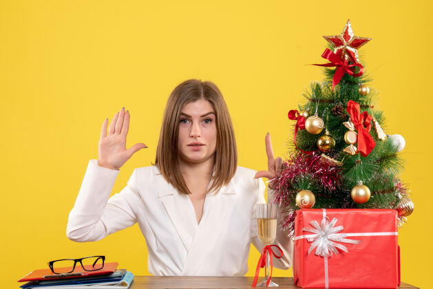女性正面图：女医生坐在黄色背景的桌子前 放着圣诞树和礼品盒人家微笑