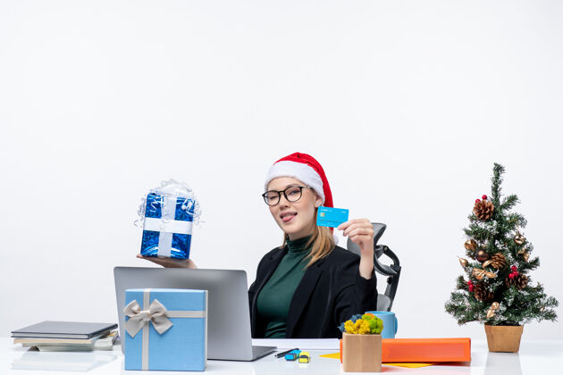 工作区戴着圣诞老人帽 戴着眼镜的女商人坐在桌子旁 手里拿着圣诞礼物和银行卡 在办公室里伸出舌头帽子圣诞老人自由职业者