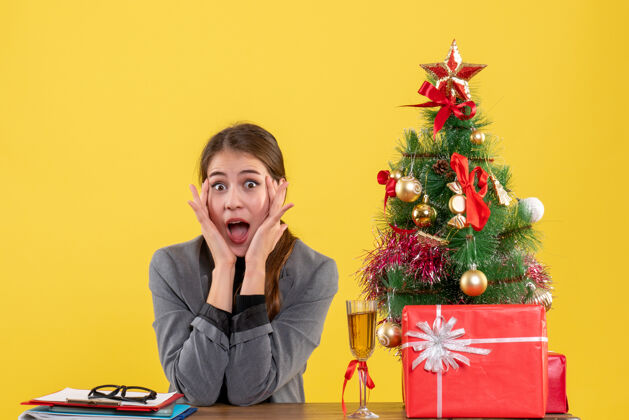 坐着前视图惊讶的女孩坐在桌子旁 把手放在脸颊上 靠近圣诞树和礼物鸡尾酒树女孩手