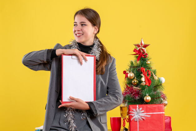 圣诞节前视图微笑的女孩拿着文件站在圣诞树和礼物鸡尾酒附近礼物桌子文件