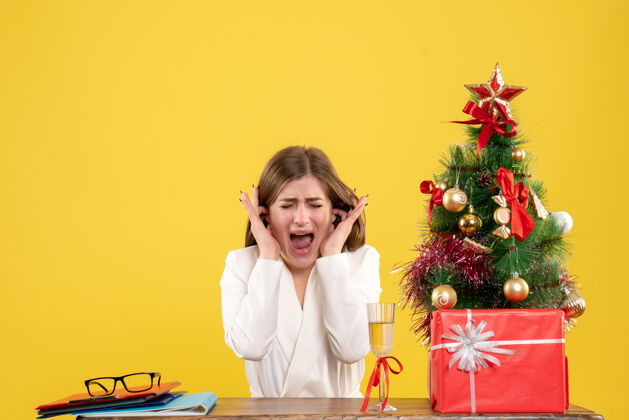 情感正面图：女医生坐在黄色背景的桌子前 放着圣诞树和礼品盒生活快乐圣诞节