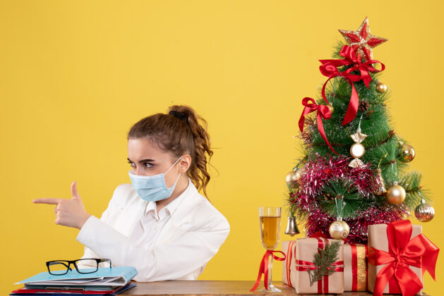 人正面图女医生戴着黄色背景的无菌口罩坐在圣诞树和礼盒里专业女性漂亮