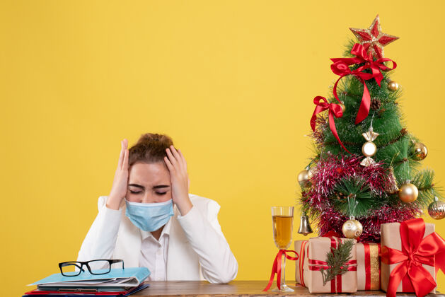 圣诞节正面图女医生戴着黄色背景的无菌口罩坐在圣诞树和礼盒里人肖像快乐