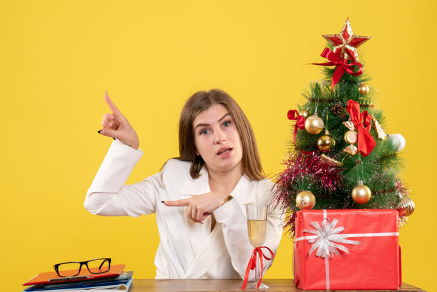 生活方式正面图：女医生坐在黄色背景的桌子前 放着圣诞树和礼品盒女医生桌子圣诞节