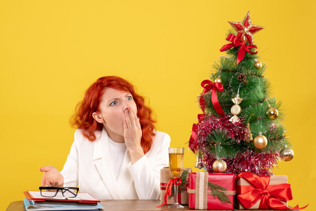 微笑前视图：女医生坐在桌子后面 黄色背景上有圣诞礼物坐着圣诞节人