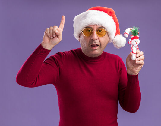 圣诞节中年男子戴着圣诞老人的黄色眼镜拿着圣诞糖果手杖看着相机惊讶地显示食指站在紫色的背景圣诞快乐相机指数