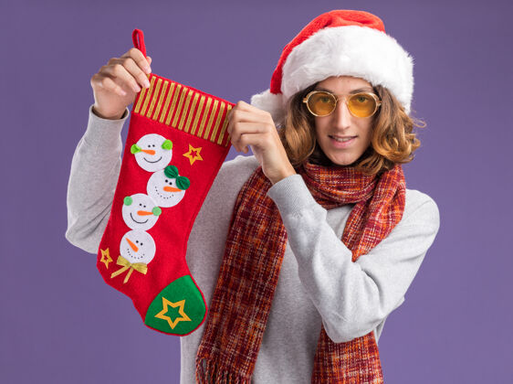 快乐快乐的年轻人戴着圣诞老人帽 戴着黄色眼镜 脖子上围着暖和的围巾 手里拿着圣诞长袜 站在紫色的背景下 面带微笑地看着相机脖子眼镜相机
