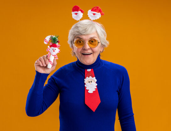 年惊讶的老妇人戴着太阳眼镜 戴着圣诞老人的头带 打着圣诞老人的领带 手里拿着一根糖果棒 背景是橙色的 还有复印空间背景新的老人