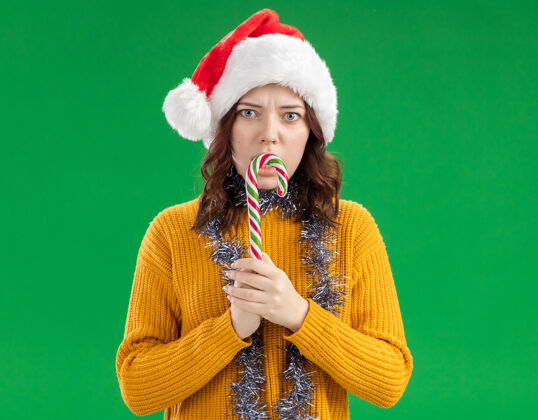 圣诞快乐焦虑的年轻斯拉夫女孩戴着圣诞帽 脖子上戴着花环 手里拿着一根被隔离在绿色背景上的糖果手杖 还有复制空间手杖焦虑年轻