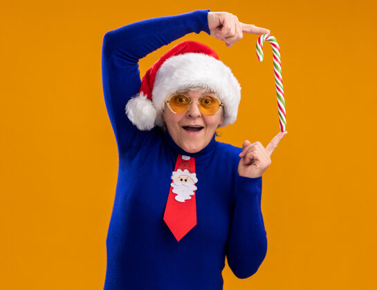 年戴着太阳眼镜 戴着圣诞帽 打着圣诞领带 拿着糖果手杖 两个手指在橙色背景上分开的老妇人惊讶不已 还有复印空间帽子领带老人