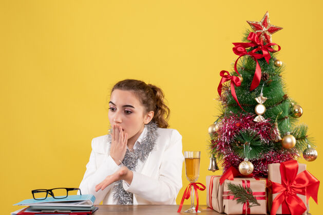 成人正面图女医生围坐在黄色背景上的圣诞礼物和圣诞树旁健康快乐圣诞树