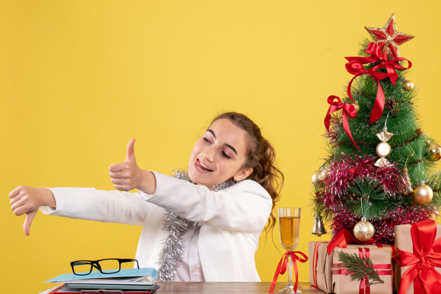 医生前视图：女医生坐在桌子后面 拿着圣诞礼物和黄色背景上的树快乐漂亮微笑