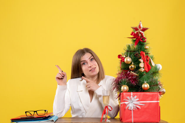 女医生前视图：女医生坐在桌子前 拿着圣诞礼物 黄色背景上的树快乐礼物微笑