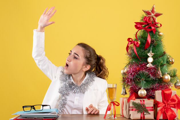 微笑前视图：女医生坐在桌子后面 拿着圣诞礼物和黄色背景上的树礼物前面护士