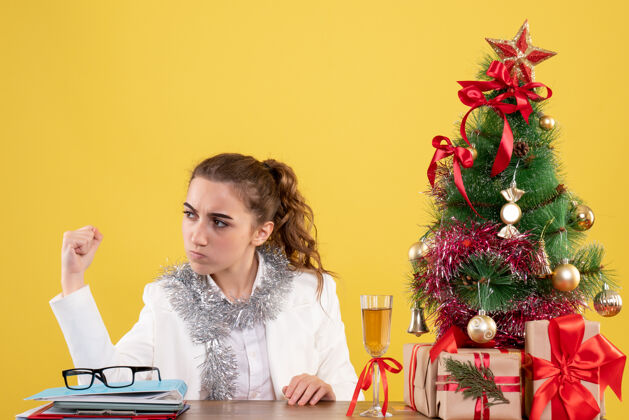 插花正面图：女医生坐在桌子后面 拿着圣诞礼物和黄色桌子上的树礼物护士树