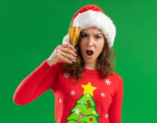 站着一个穿着红色圣诞毛衣 戴着圣诞帽的年轻女人 手里拿着一杯香槟 站在绿色的背景下 看着摄像机 既恼火又恼火帽子年轻杯子